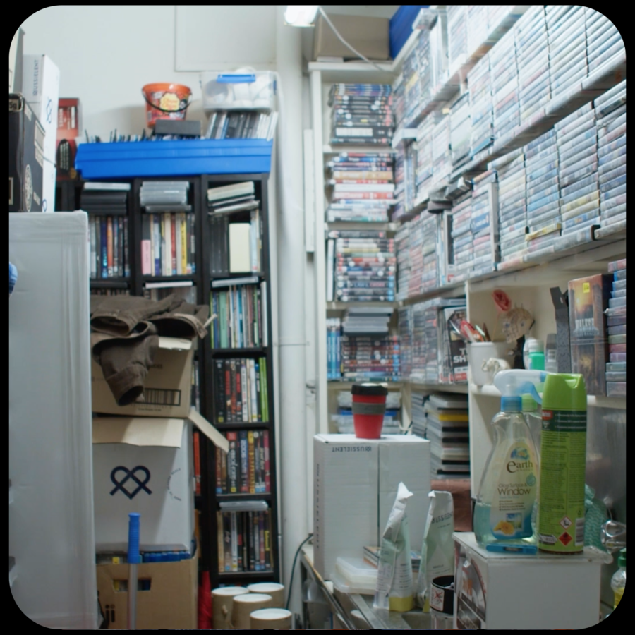 A behind the scenes view of a video shop store room, with shelving almost to the ceiling full of DVDs and assorted storage boxes, cleaning products and coffee mugs amongst them.